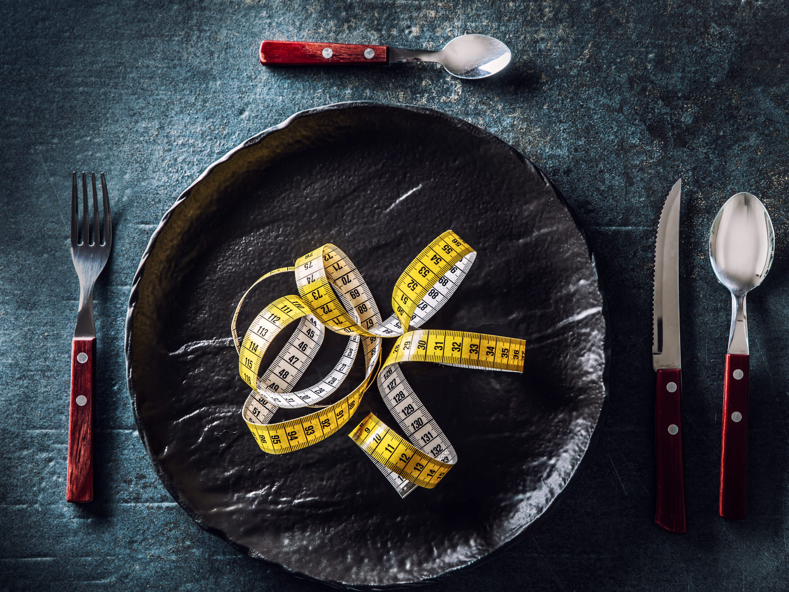 Dark plate with measure tape, next to it cutlery. Top of view on concept healthy eating.