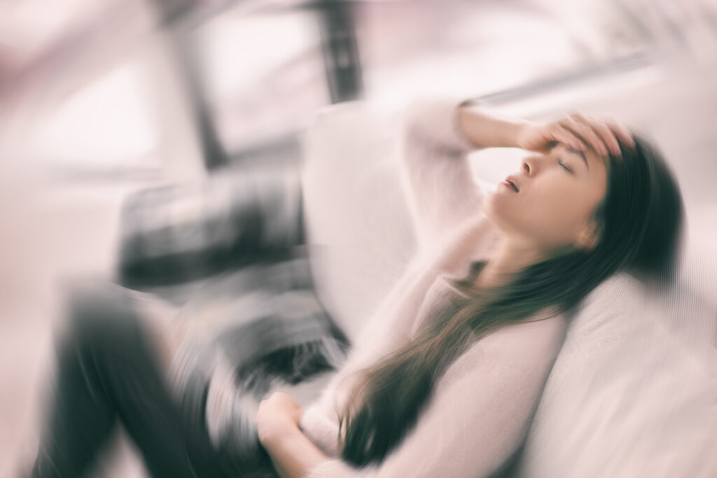 A woman sitting on a couch with a blurred, dizzy background effect, holding her head and stomach, appearing exhausted and overwhelmed - representing chronic fatigue or burnout