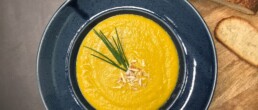 A bowl of smooth carrot and orange soup served in a deep blue ceramic plate. The bowl is placed on a rustic wooden board alongside a slice of toasted sourdough and a loaf of crusty bread. The background is a textured white surface, creating a cozy, artisanal presentation.
