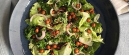 A vibrant salad made of kale, romaine lettuce, roasted chickpeas, and slivered red onions, garnished with sesame seeds and chilli flakes. A light cashew dressing is drizzled over the greens.