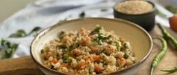 A close-up of a rustic ceramic bowl filled with quinoa upma, featuring vibrant chopped carrots, green peas, French beans, and garnished with fresh coriander. The bowl rests on a wooden board, surrounded by green chilies, raw quinoa in a small bowl, and fresh vegetables, creating a warm and inviting food presentation.