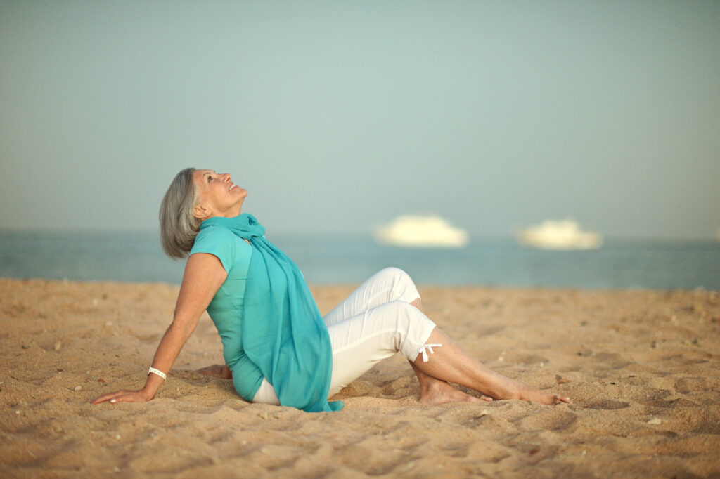 An older woman sitting relaxed on a sandy beach, leaning back and looking up peacefully, with the ocean and blurred boats in the background.