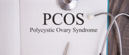 Open notebook with the words “PCOS Polycystic Ovary Syndrome” written on a grid page, accompanied by a stethoscope and metal pen on a wooden surface