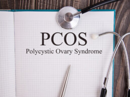 Open notebook with the words “PCOS Polycystic Ovary Syndrome” written on a grid page, accompanied by a stethoscope and metal pen on a wooden surface