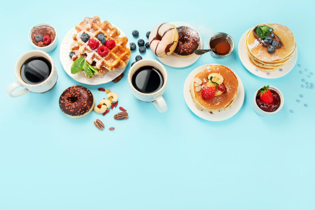 An assortment of sweet breakfast items on a light blue background, including waffles with berries, pancakes with fruit, donuts, chocolate, syrup, nuts, and three cups of black coffee.