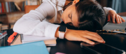 Tired woman asleep at her desk, surrounded by notebooks, a laptop, and a smartphone - reflecting work fatigue and lack of proper rest