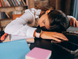 Tired woman asleep at her desk, surrounded by notebooks, a laptop, and a smartphone - reflecting work fatigue and lack of proper rest
