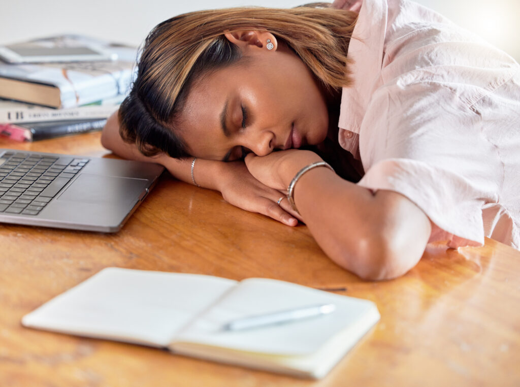 A tired woman with her head resting on a wooden desk, asleep beside an open laptop and notebook, suggesting fatigue or burnout.